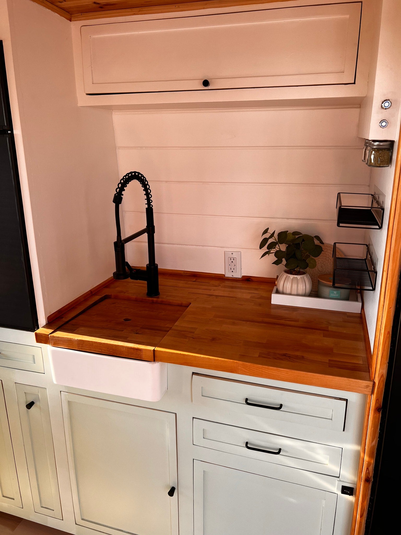 This modern RV rental kitchenette features light wood countertops, a white farmhouse sink, and a black faucet, with pale green cabinets below.