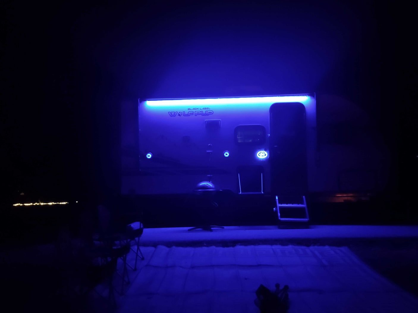 A modern Wolf Pup RV rental trailer illuminated in vivid blue lights at night, with steps and a mat outside.