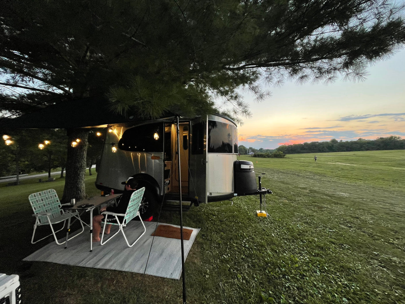Unwind on your RV rental adventure! This image shows a camper, outdoor seating, and string lights at sunset by a field.