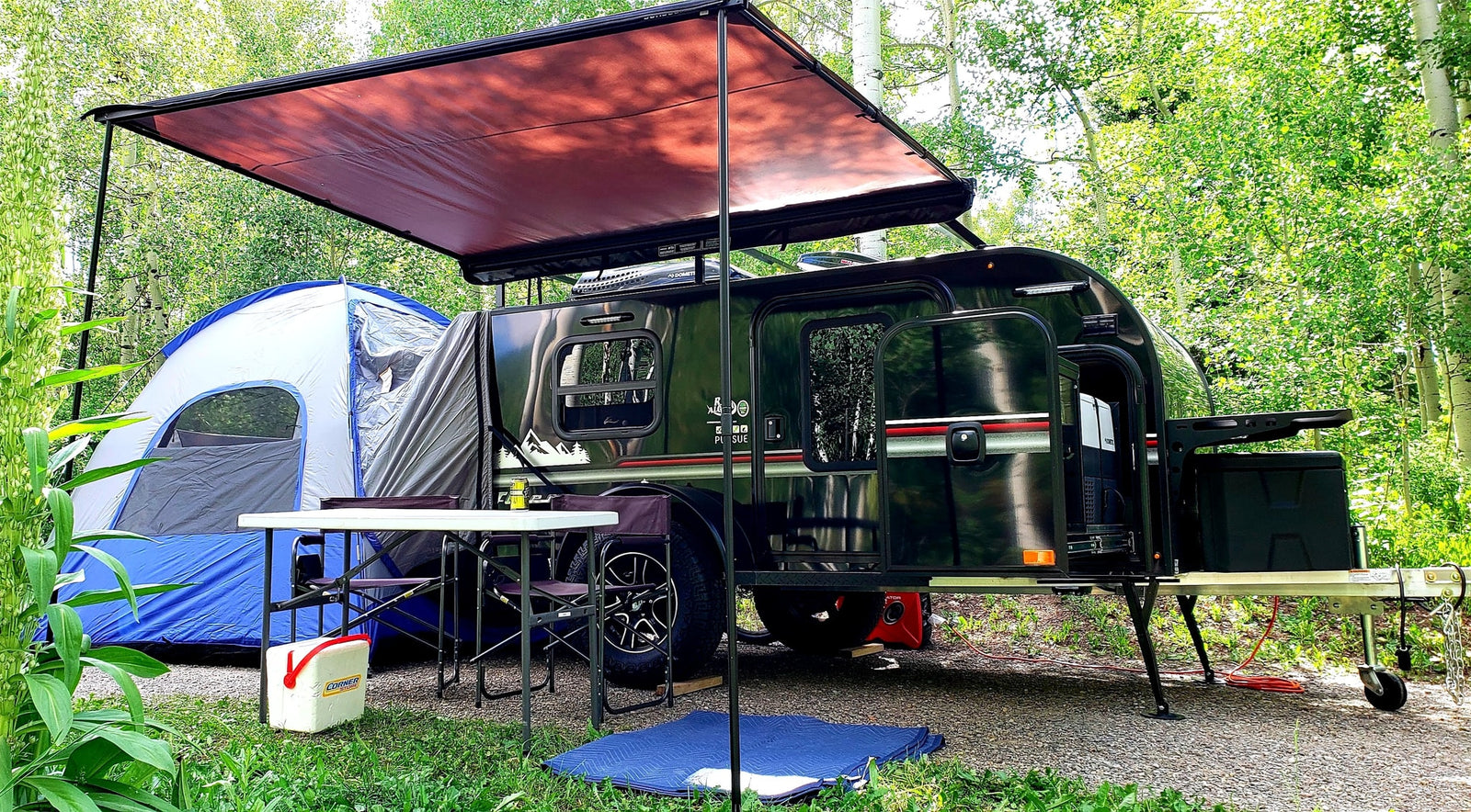 Scenic rv rental campsite: dark teardrop trailer, extended awning, blue tent, and outdoor table setup amidst green trees.