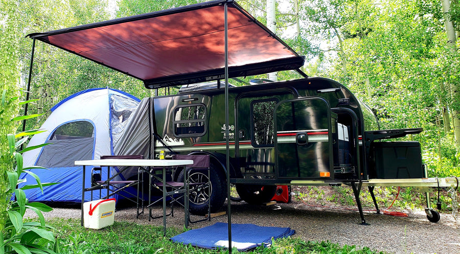 Scenic rv rental campsite: dark teardrop trailer, extended awning, blue tent, and outdoor table setup amidst green trees.