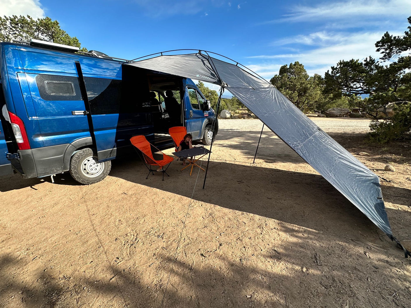 An RV rental camper van with an extended awning provides shade for chairs & a child on a sunny day.