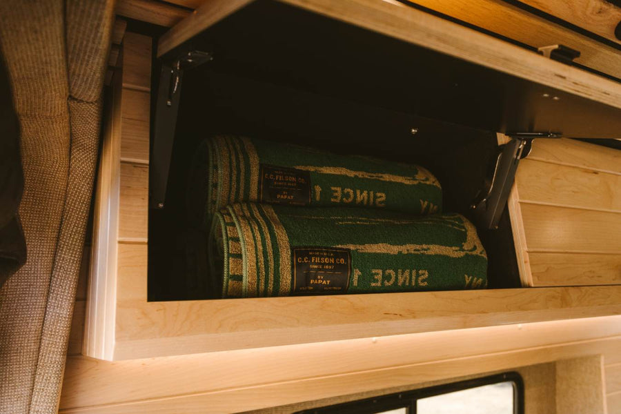 Organized overhead cabinet in an RV rental, featuring two green C.C. Filson blankets and warm wooden finishes.