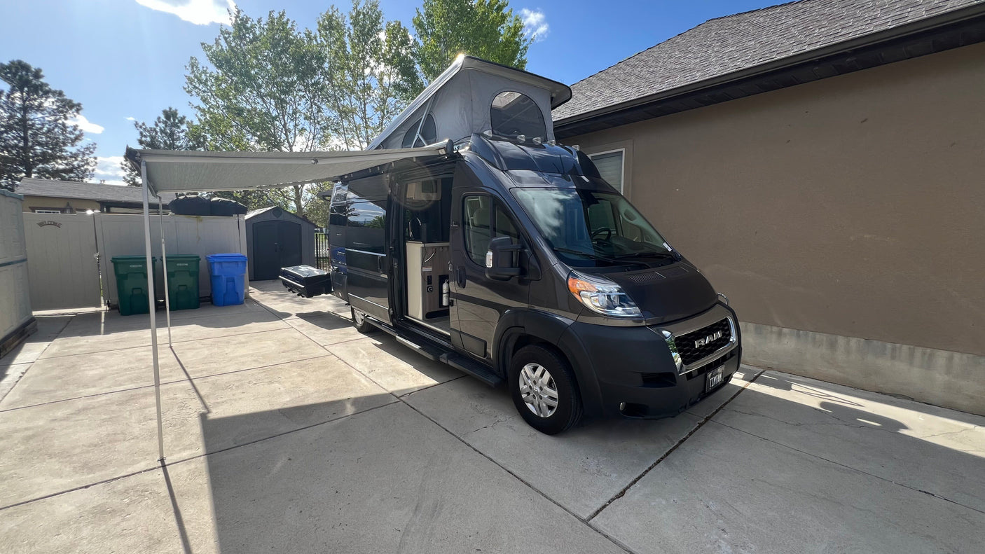 This dark gray camper van, with its awning extended and pop-top raised, is ready for your next RV rental adventure.