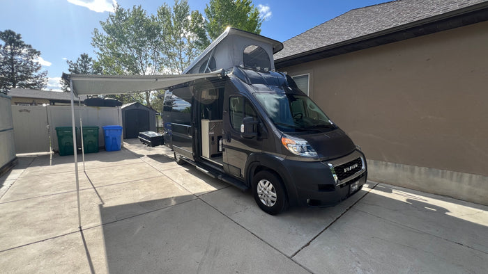 This dark gray camper van, with its awning extended and pop-top raised, is ready for your next RV rental adventure.