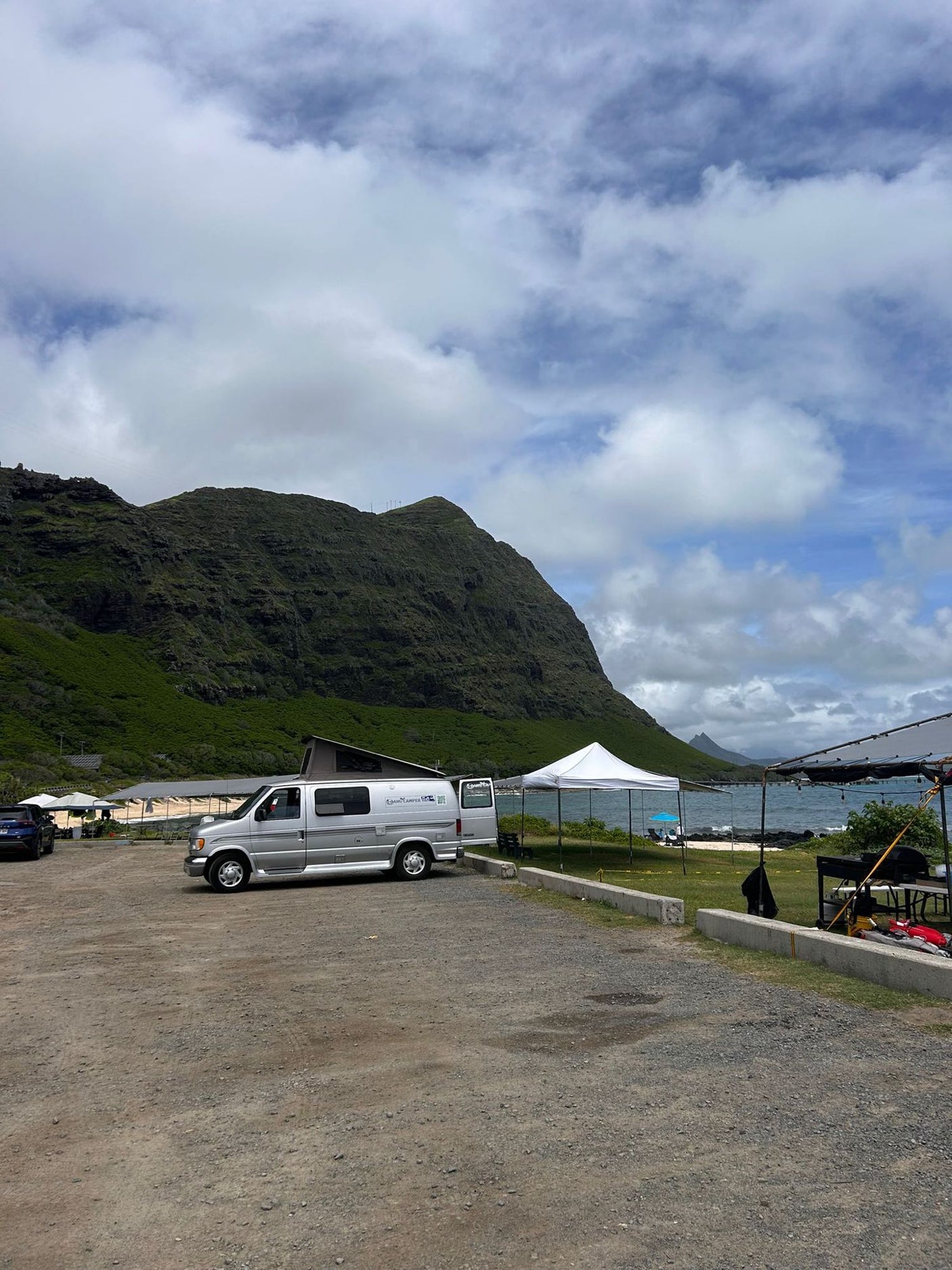 A silver RV rental van is parked on a gravel lot, with a lush green mountain behind and the ocean shoreline to the right. Outdoor tents are visible.