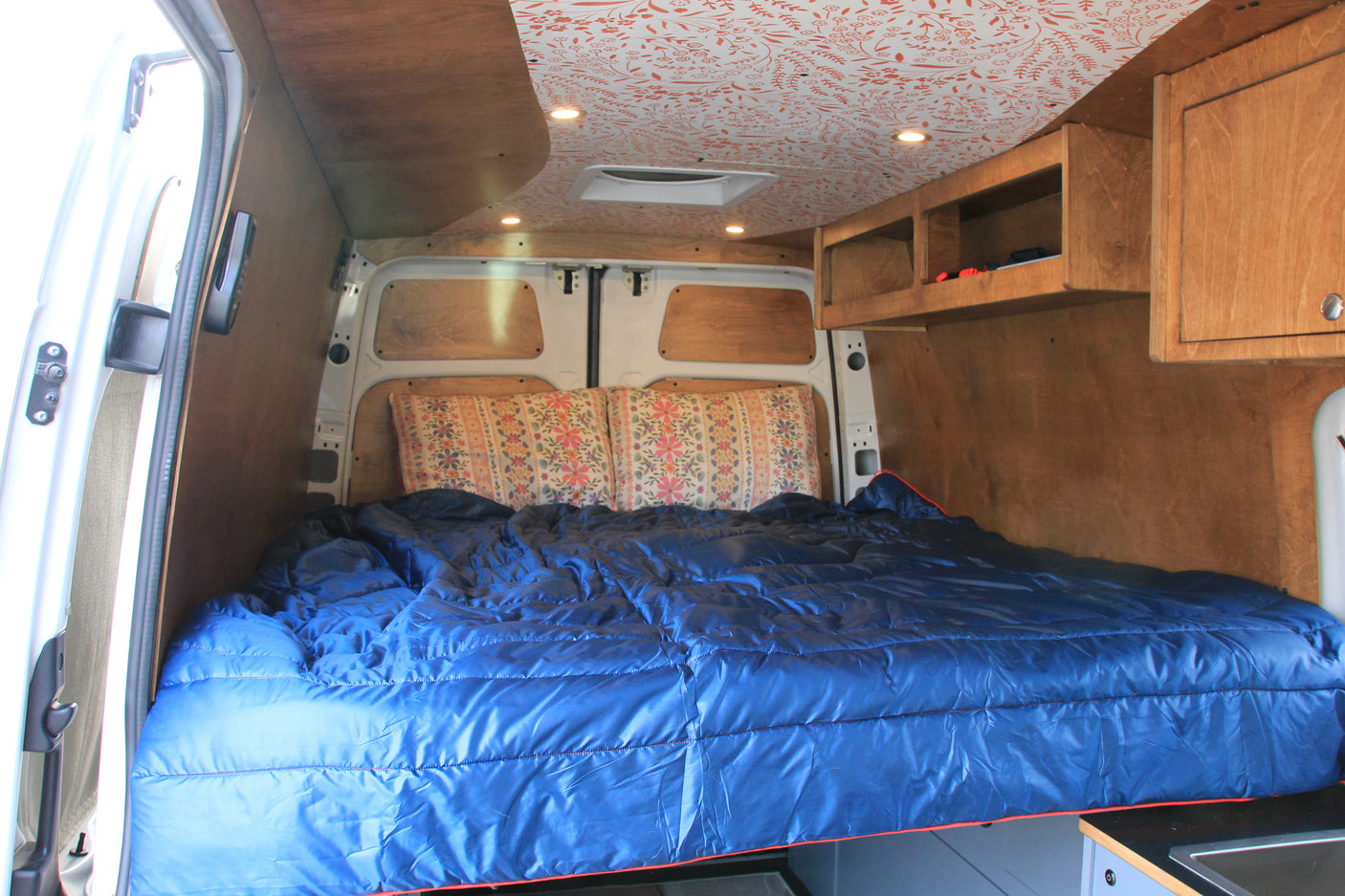 Inside an RV rental: Custom campervan interior featuring a bed with blue bedding, wood walls, cabinets, and a patterned ceiling.