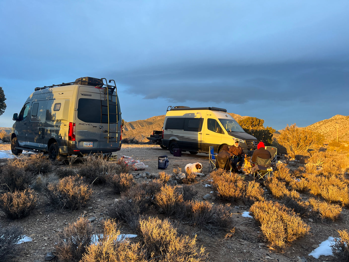 Two Winnebago Revel RV rental vans set up for boondocking. Campers and a dog gather around a warm campfire at sunset.