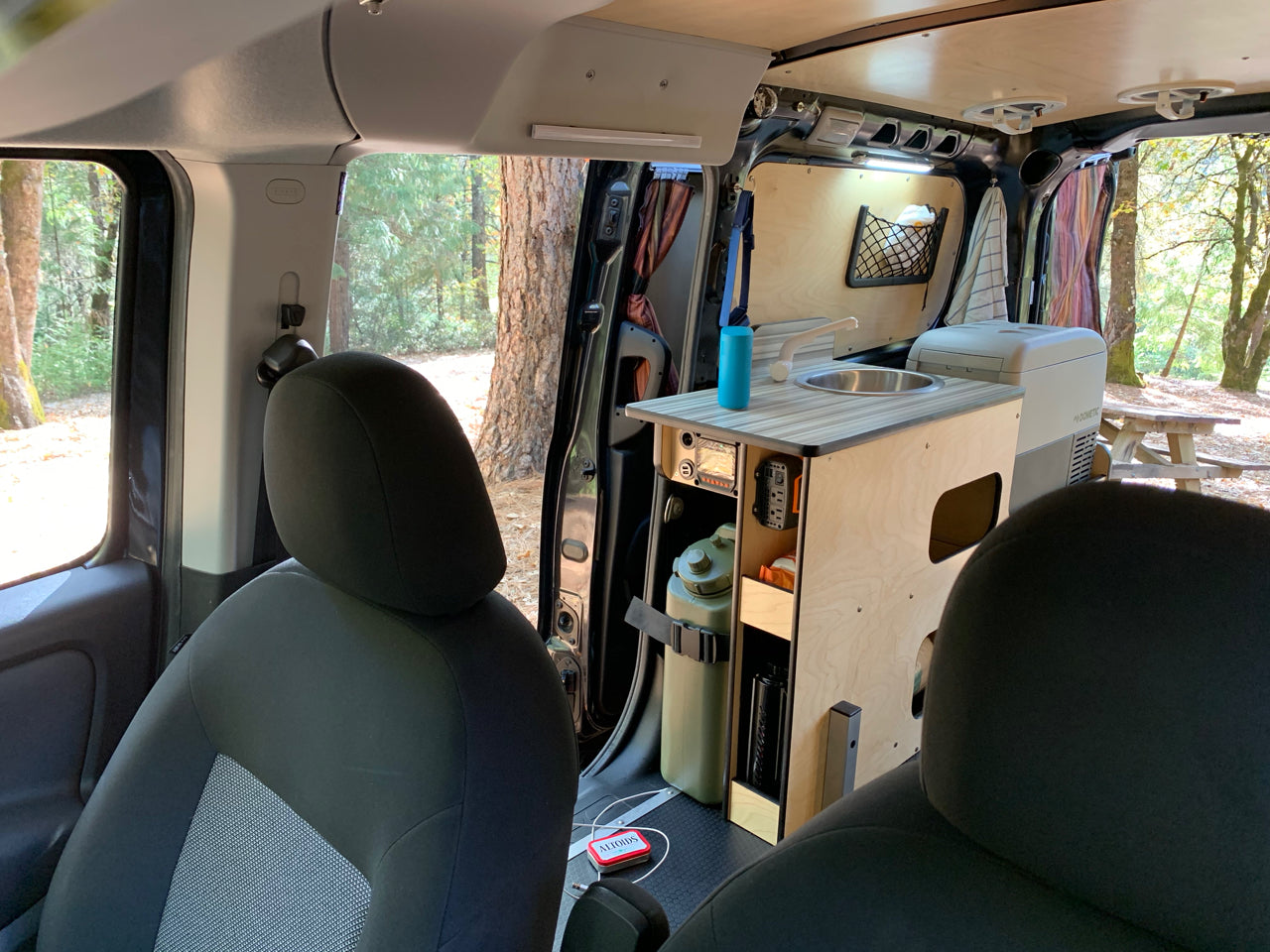 Compact RV rental interior featuring a custom kitchenette with sink, Dometic fridge, and built-in storage for travel adventures among trees.