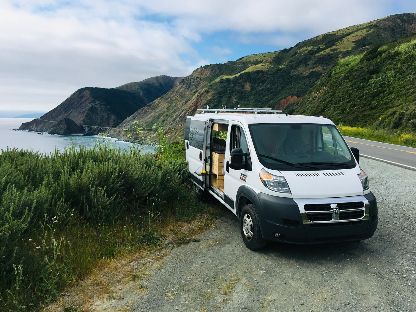 White RV rental van at a stunning coastal overlook with the iconic Bixby Bridge and Pacific Ocean in California.|Stylish RV rental conversion showing a cozy bed — photo 1