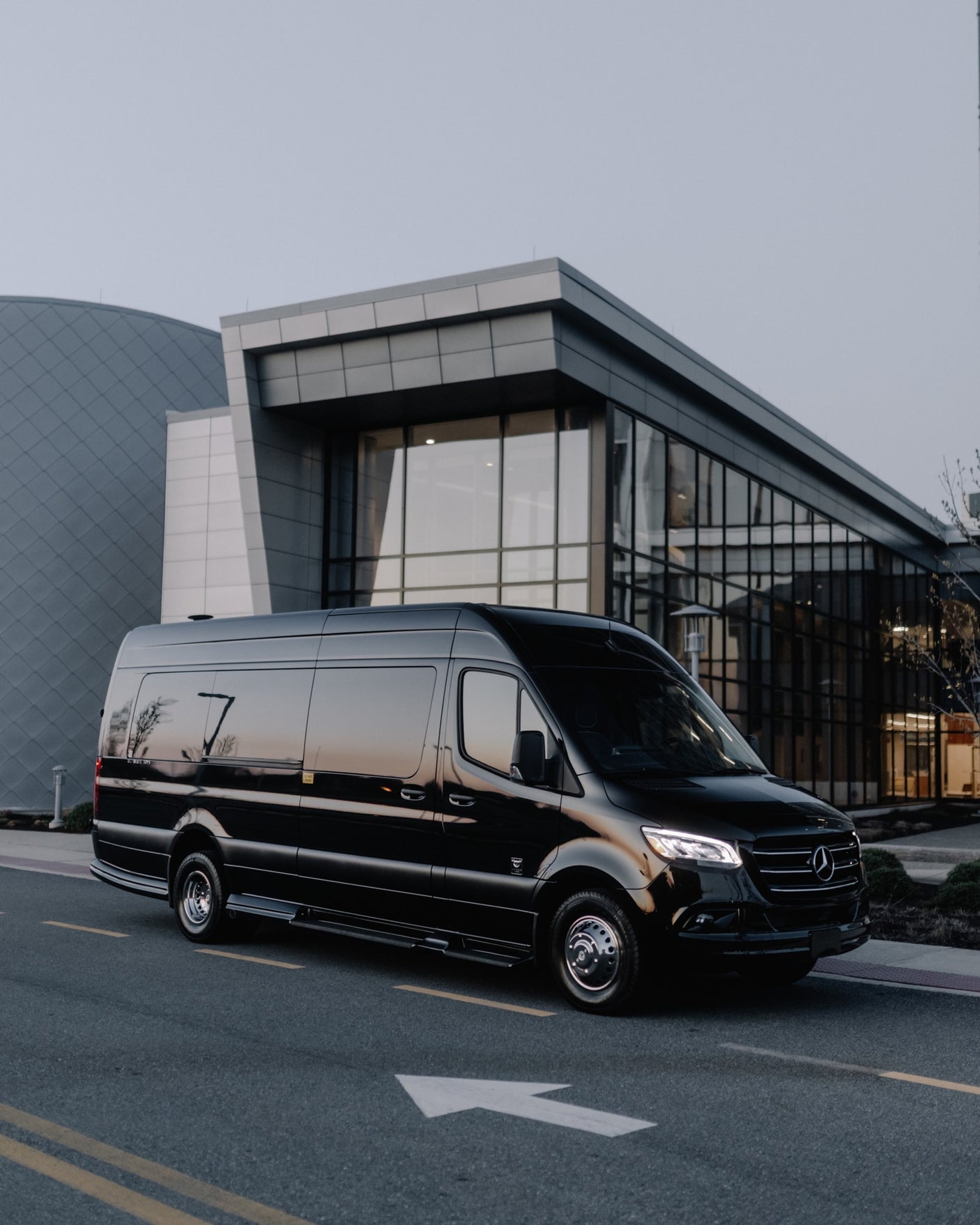 Sleek black Mercedes-Benz Sprinter luxury van for RV rental, parked on road at dusk with modern buildings in background.