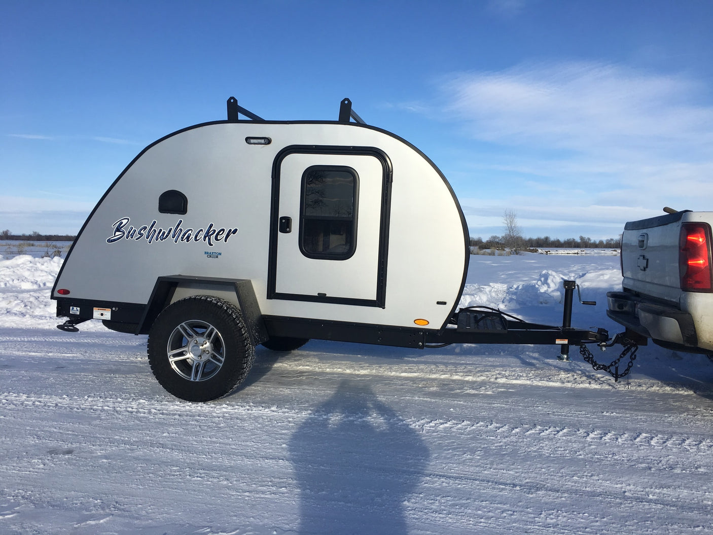 White Bushwhacker teardrop RV rental hitched to a truck on a snowy landscape under a clear blue sky.