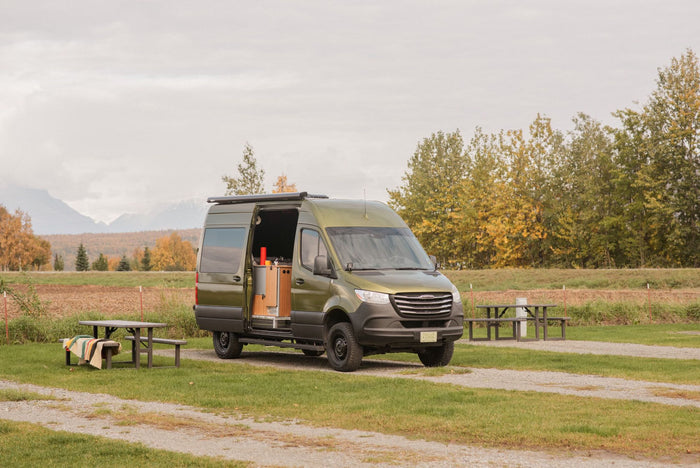 set against snow-capped mountains.|Green camper van RV rental parked at a scenic campsite with mountains & fall trees.|Green camper van RV rental parked at a mountain campsite with an open side door and picnic table.|Green RV rental camper van at an autumn campsite