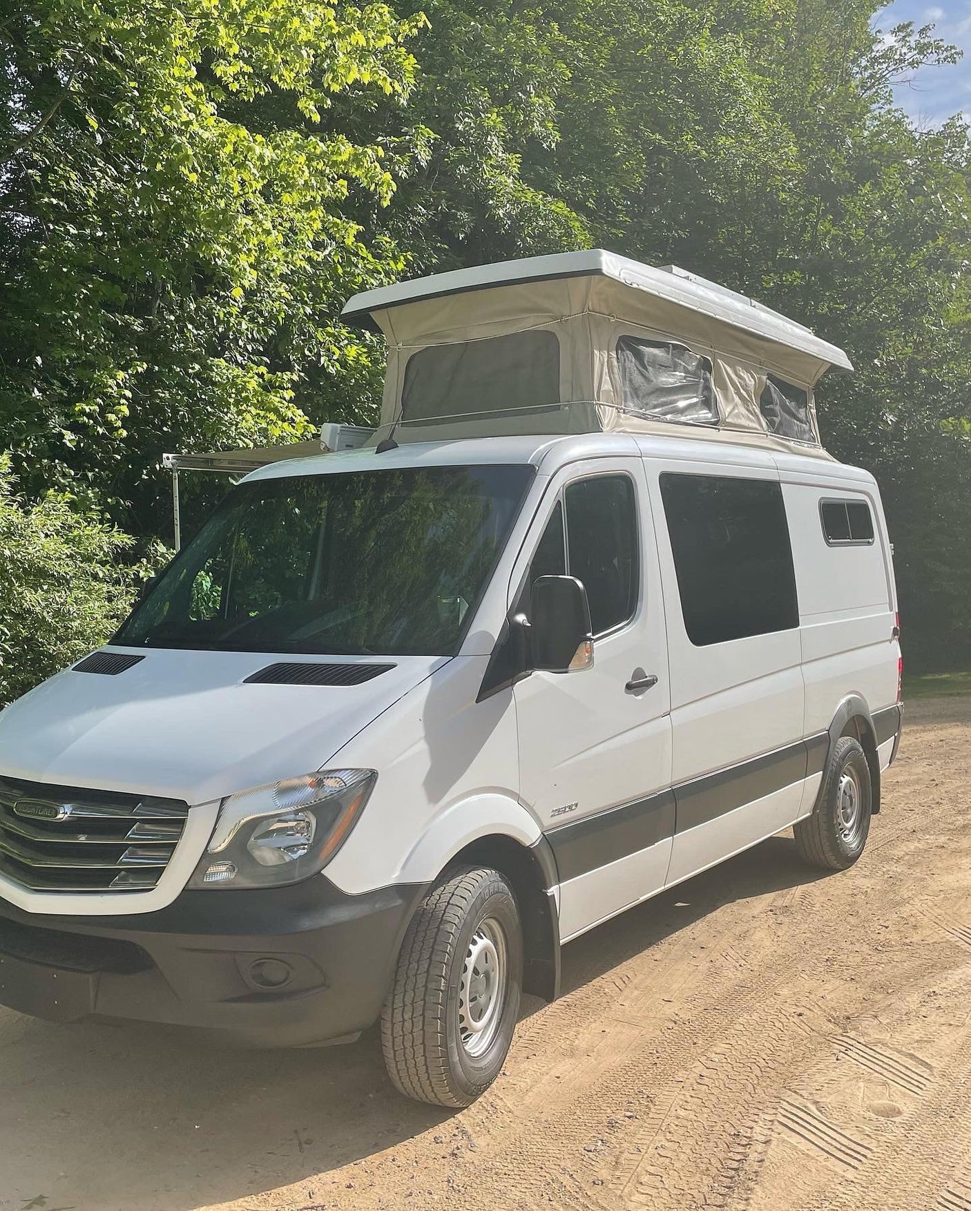 is parked on a dirt road in a sunny forest.|Cozy pop-top RV rental sleeping area with plush mattress