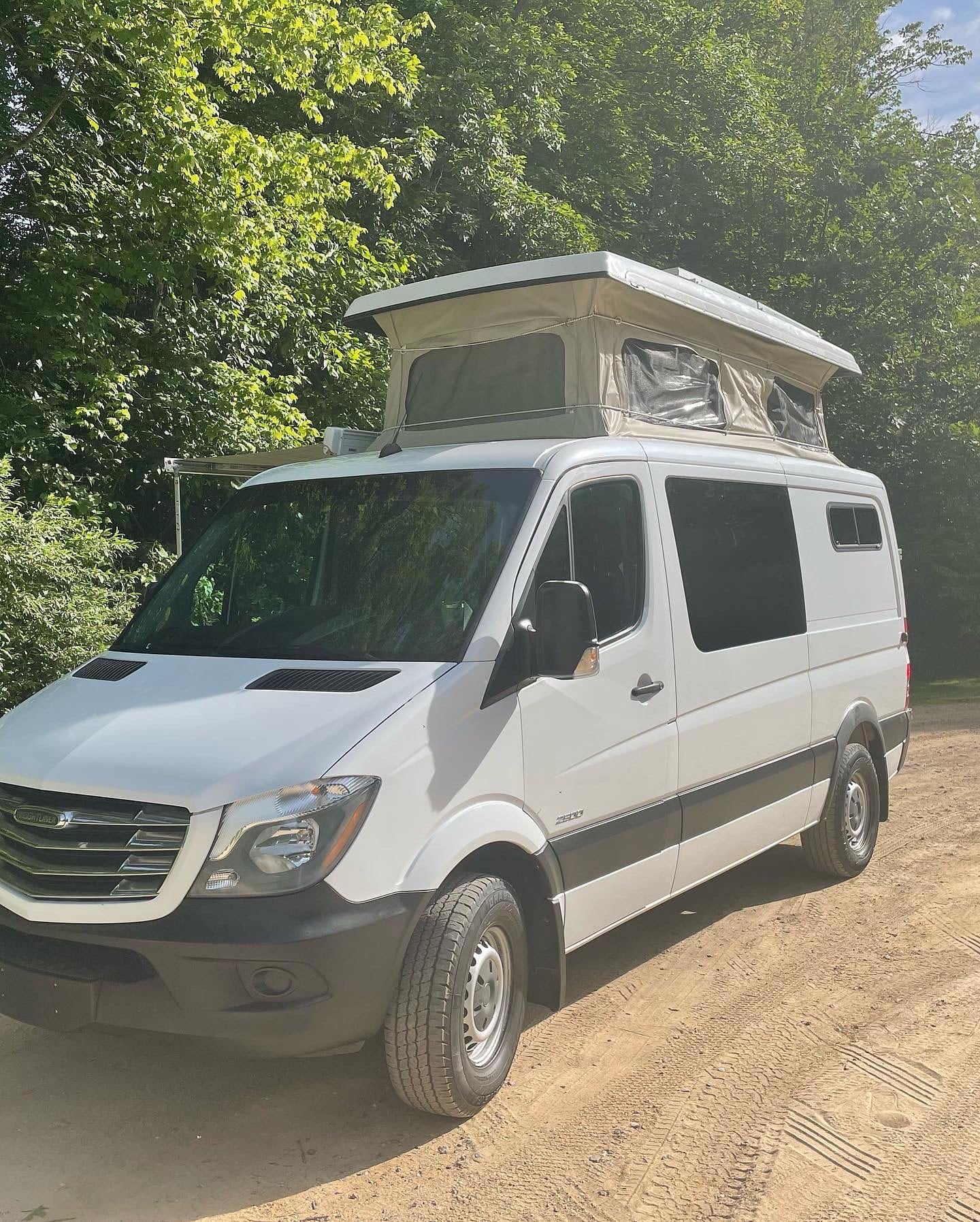 is parked on a dirt road in a sunny forest.|Cozy pop-top RV rental sleeping area with plush mattress