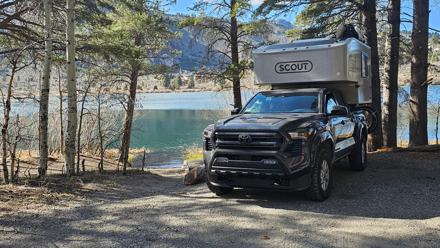 Explore nature with this truck camper, an ideal RV rental. Parked lakeside, surrounded by trees and mountains, ready for adventure.