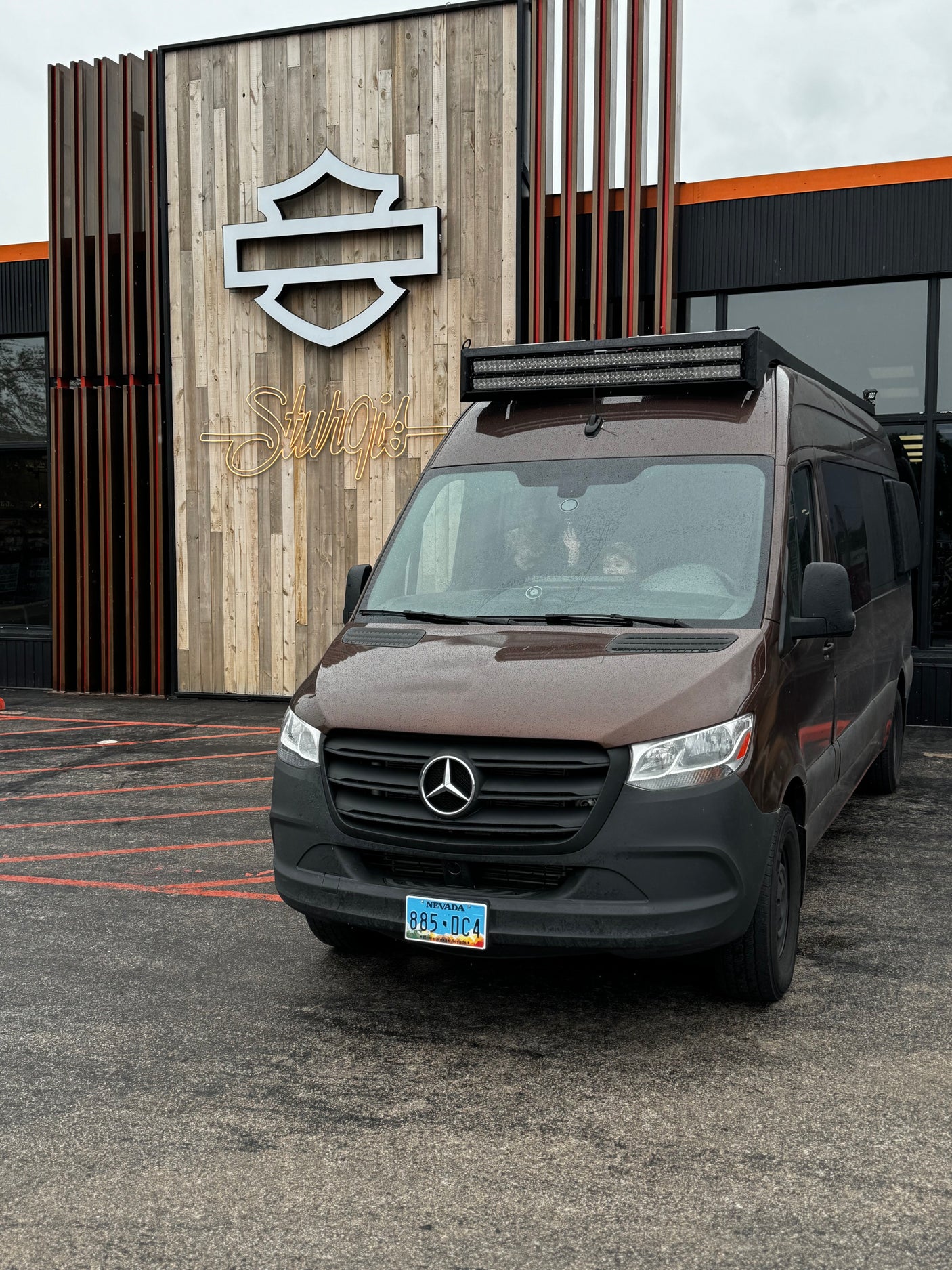 Brown Mercedes-Benz Sprinter rv rental van with a roof light bar, parked outside a Harley-Davidson Sturgis building.