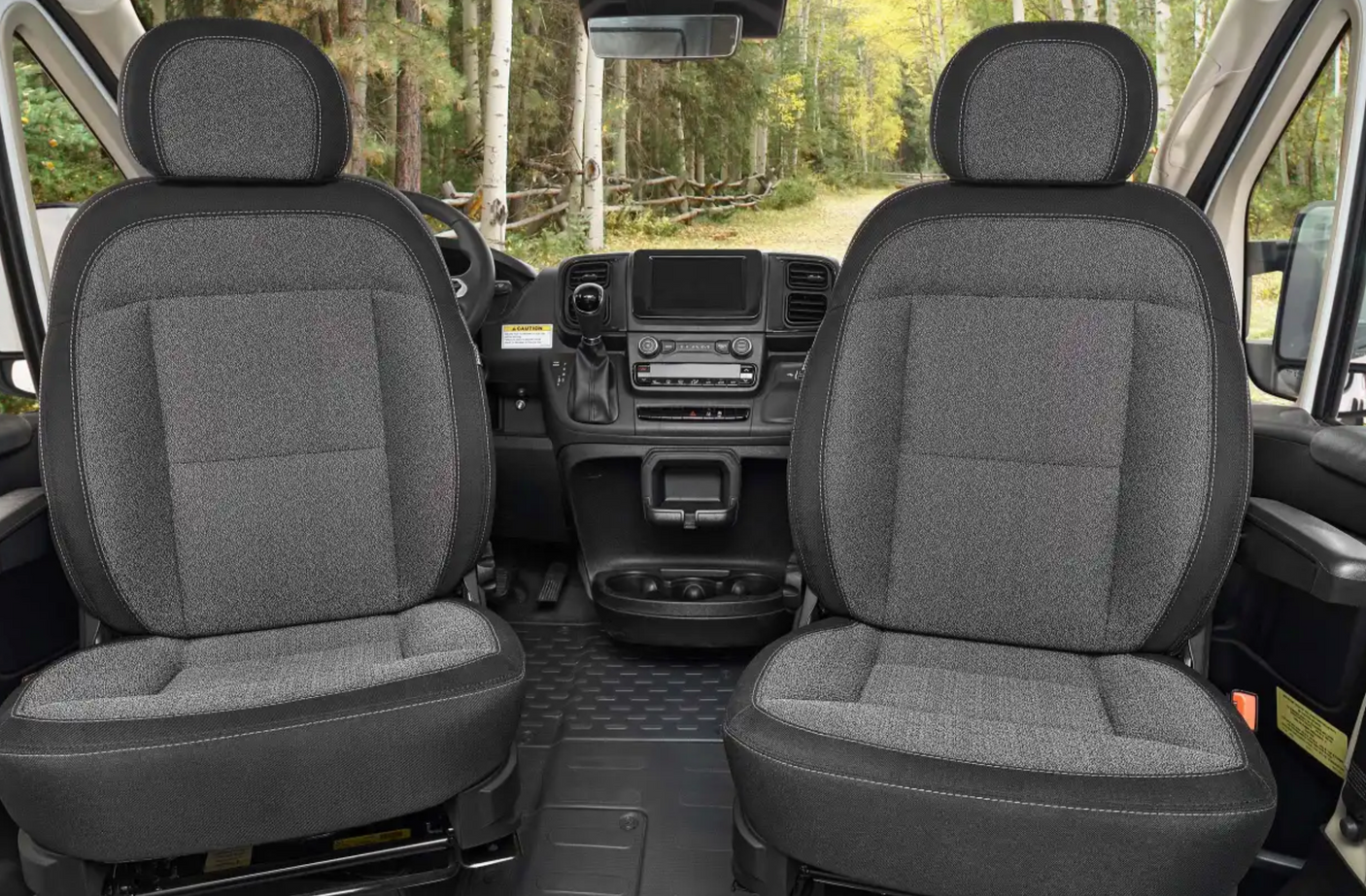 Spacious RV rental cabin featuring comfortable dark grey fabric seats, a modern dashboard, and a view of a forested road.