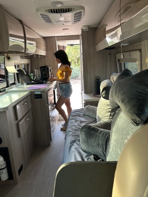 A woman works in the kitchen of a modern RV rental, featuring a comfortable bed & open door to a natural setting.