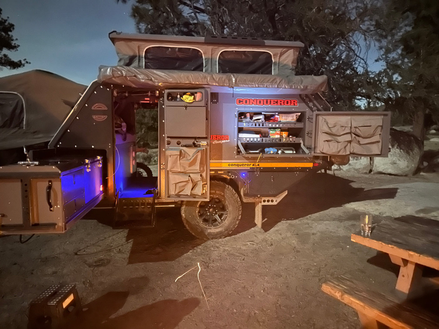 This illuminated Conqueror off-road camper, perfect for RV rental trips, reveals its outdoor kitchen and rooftop tent setup at night.