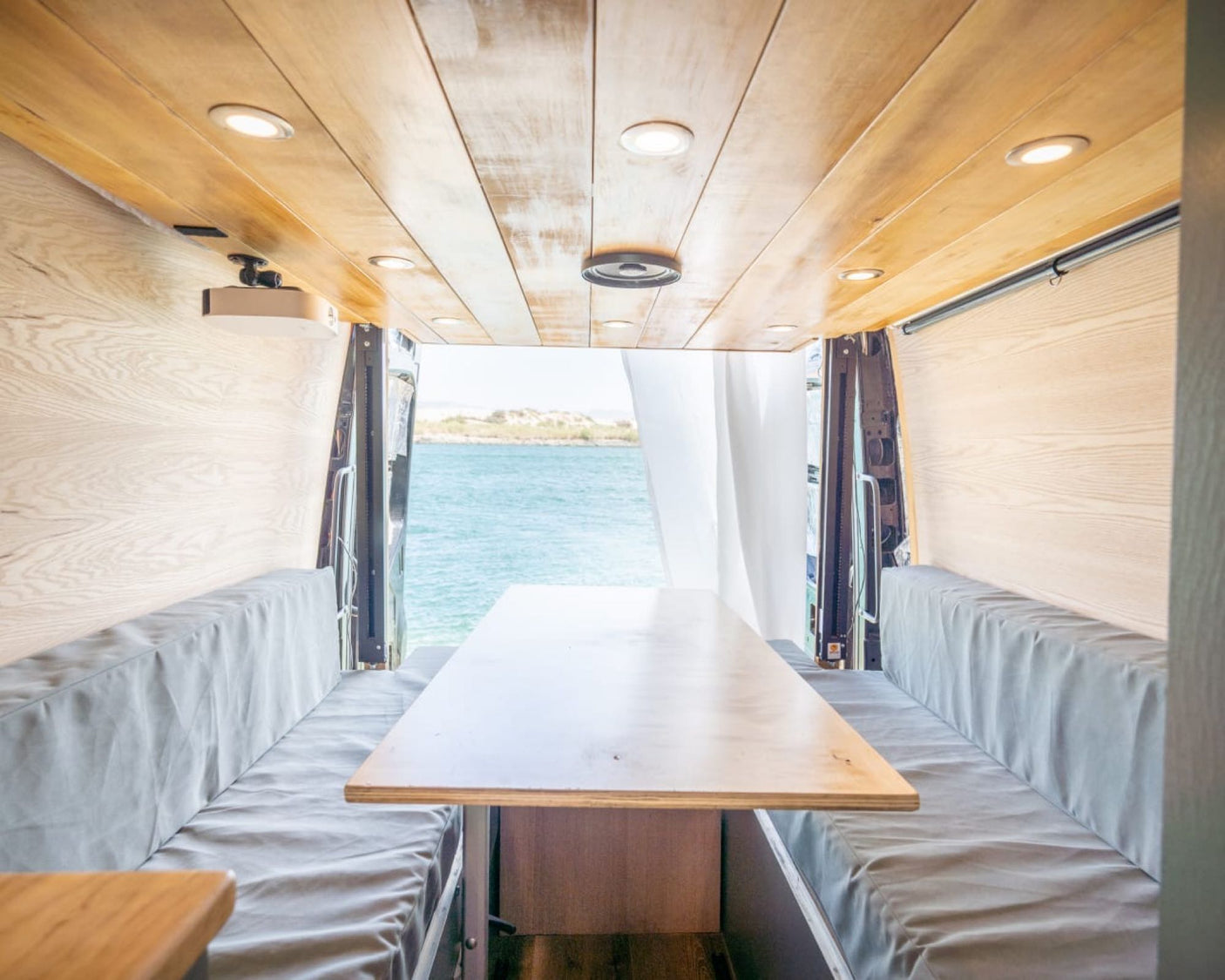 Modern rv rental camper interior: light wood, dining table, benches, and bright ocean view from open back doors.