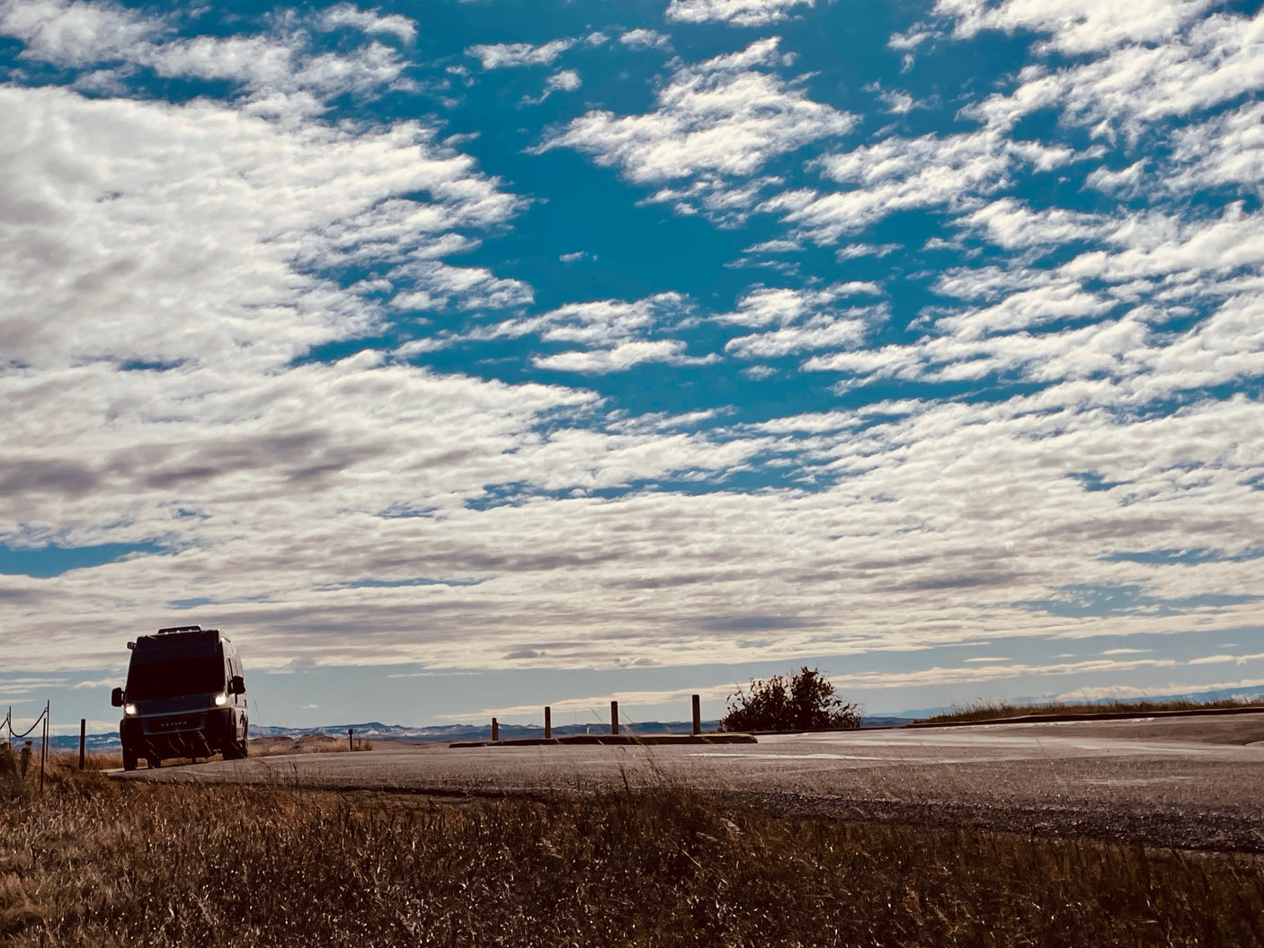 Open road freedom! A dark RV rental van drives on a rural road under an expansive blue sky full of white clouds.