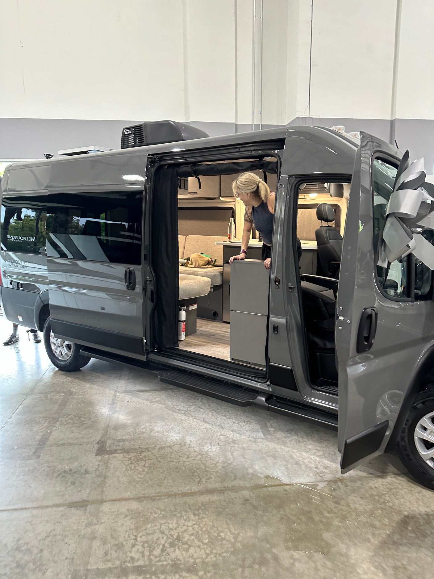 A woman inspects the interior of a new gray camper van, adorned with a large bow, ready for an RV rental.