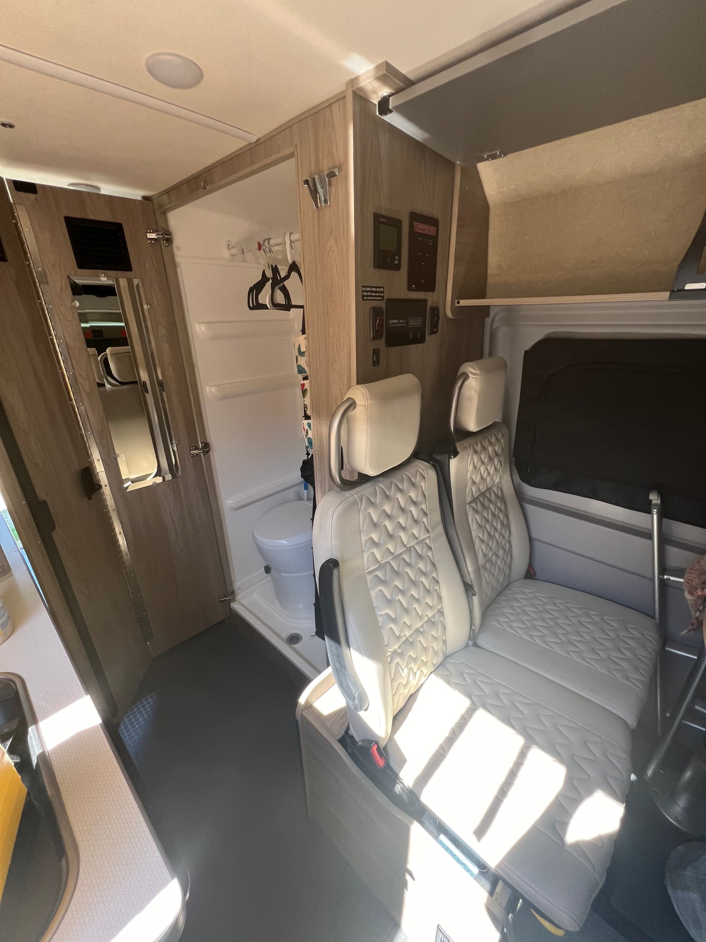 Bright interior of an RV rental, featuring cream quilted seats, a compact wet bath with toilet, and control panels.