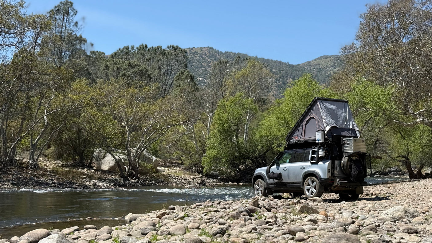 Overland-equipped SUV with rooftop tent on a rocky riverbank, surrounded by trees and mountains. An ideal RV rental for nature adventures.