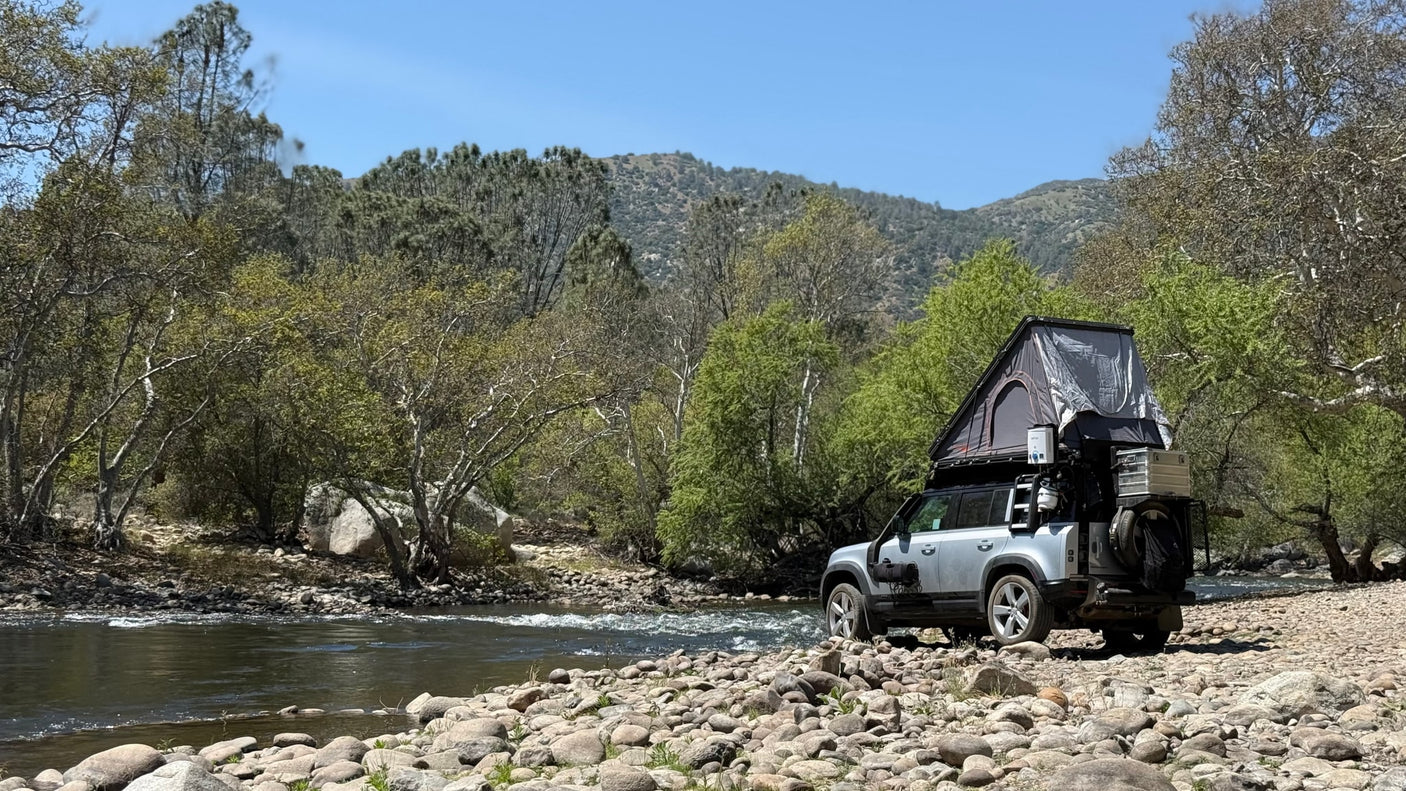 Overland-equipped SUV with rooftop tent on a rocky riverbank, surrounded by trees and mountains. An ideal RV rental for nature adventures.