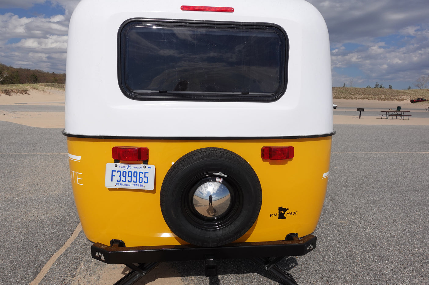 Yellow teardrop RV rental parked near sandy dunes. This MN-made trailer with a Michigan plate features a rear spare tire.