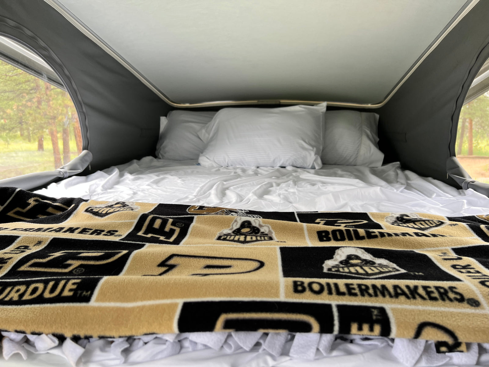Inside an RV rental pop-top: a comfy bed with white linens and a Purdue Boilermakers blanket, surrounded by nature views.