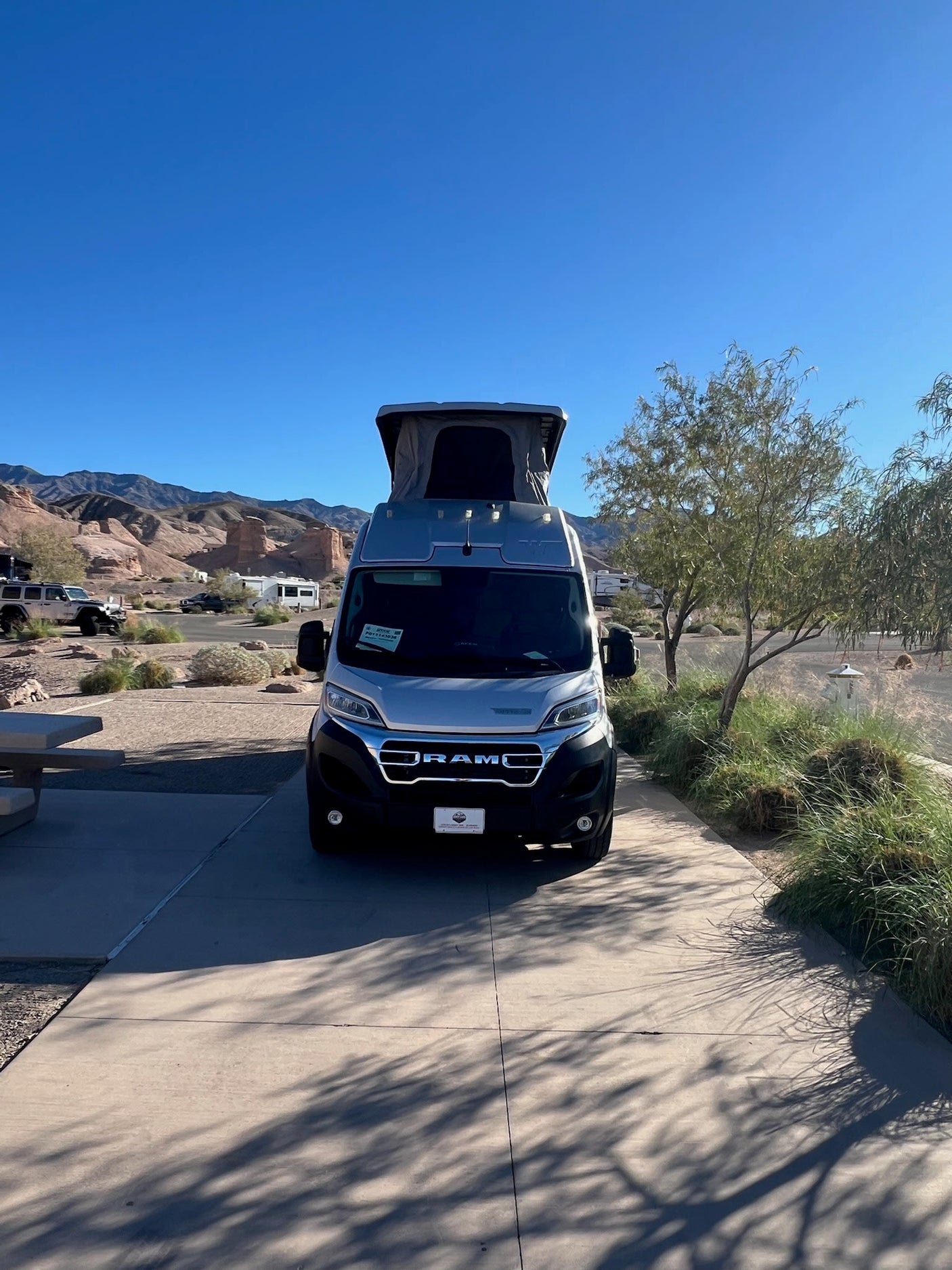 Front view of a silver RV rental with a pop-top at a scenic desert campsite. Mountains and clear blue skies abound.