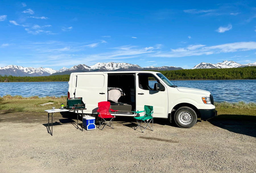 White RV rental van parked lakeside with majestic snow-capped mountains. Complete with camp setup: stove
