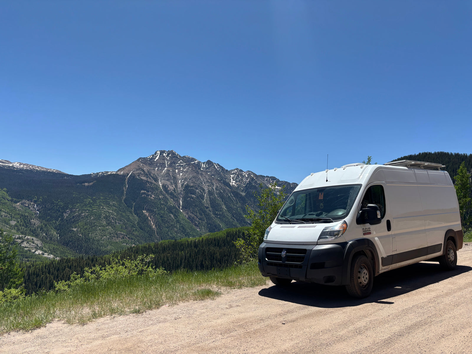 This RV rental, a white camper van, enjoys a scenic mountain view with snow-capped peaks and lush forests.