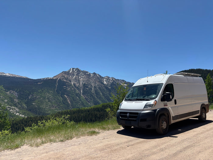 This RV rental, a white camper van, enjoys a scenic mountain view with snow-capped peaks and lush forests.