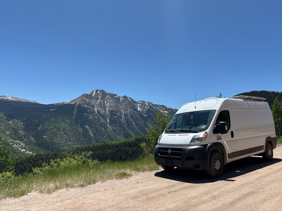 This RV rental, a white camper van, enjoys a scenic mountain view with snow-capped peaks and lush forests.