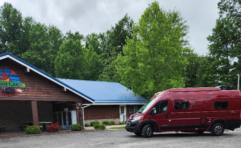 A shiny red rv rental camper van parked outside a rustic lodge with a blue roof, surrounded by lush green trees.