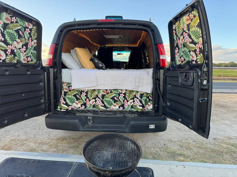 Inside an open **rv rental** camper van: bed with tropical decor, string lights, and grill setup for outdoor adventure.