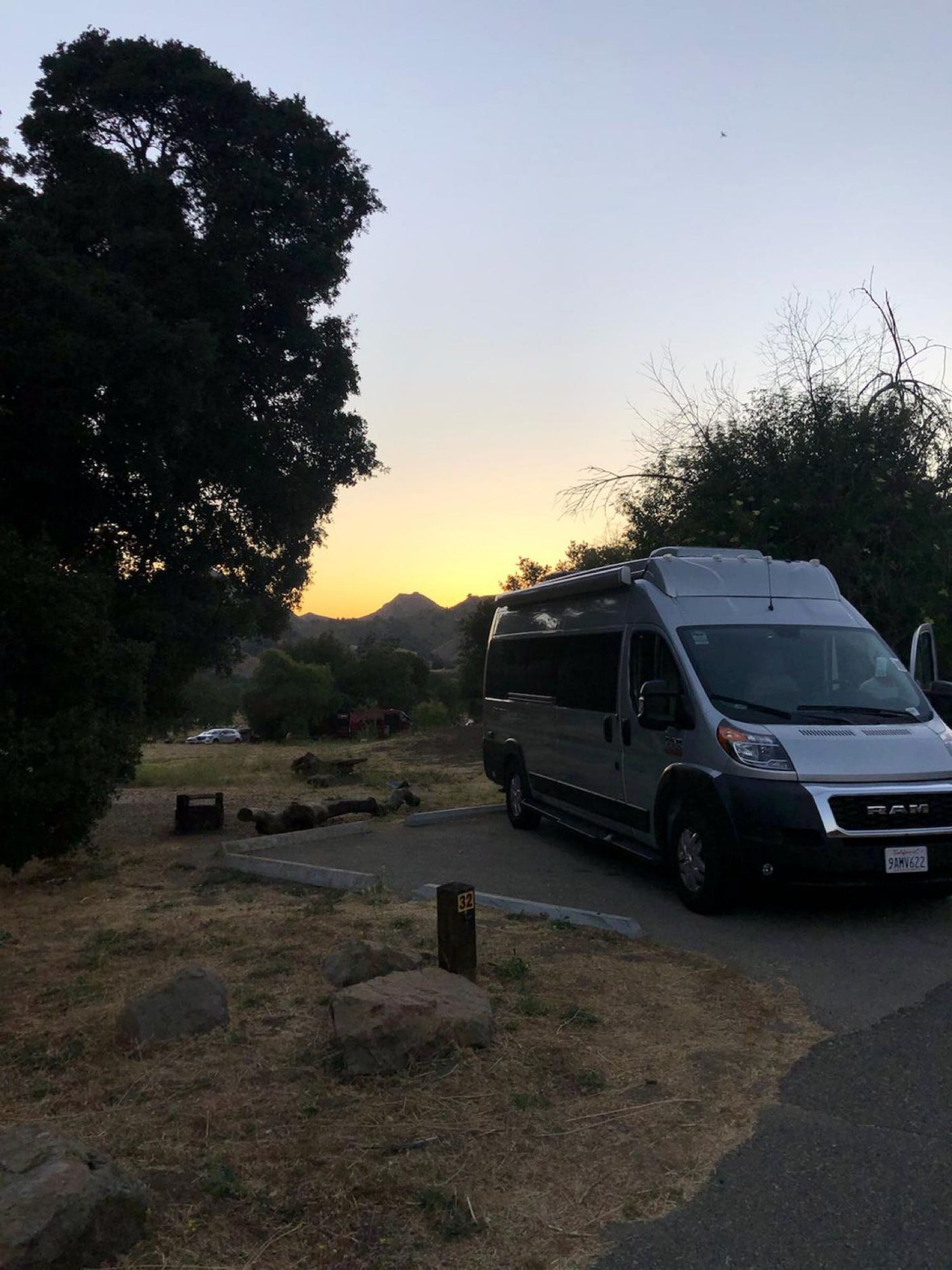 A grey RV rental parked in campsite #32, with a golden sunset behind distant mountains and trees.