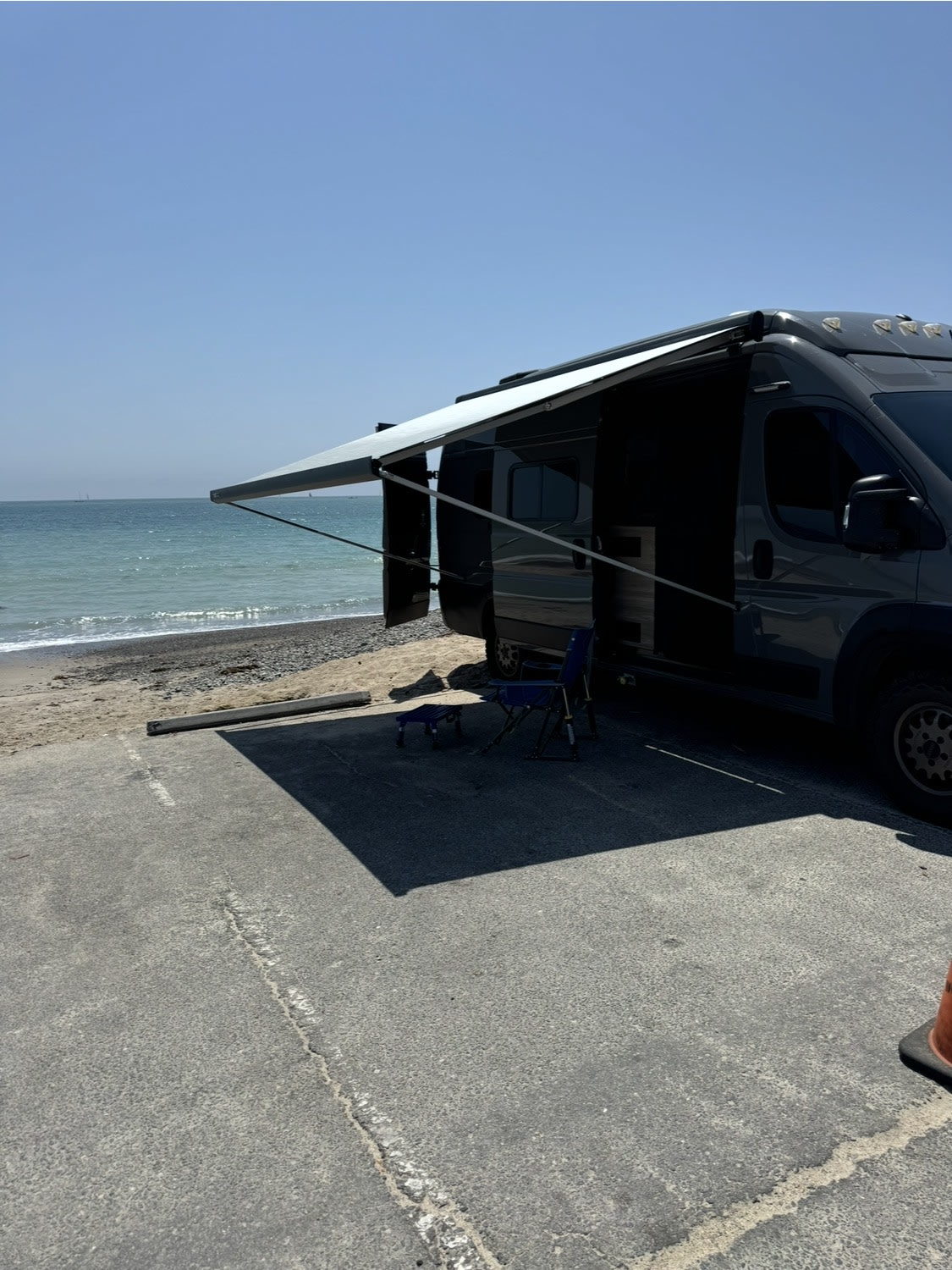 parked by the road.|RV rental camper van with pop-top roof and blue tent at a sunny forest campsite. A large rock is nearby on the dirt ground.|This grey Ram ProMaster rv rental — photo 3