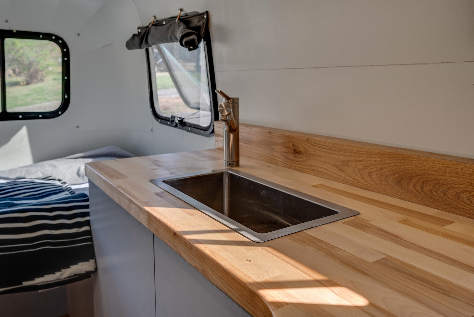 Inside an RV rental: a compact kitchen with a light wood countertop, stainless steel sink, and modern faucet, beside a bed.