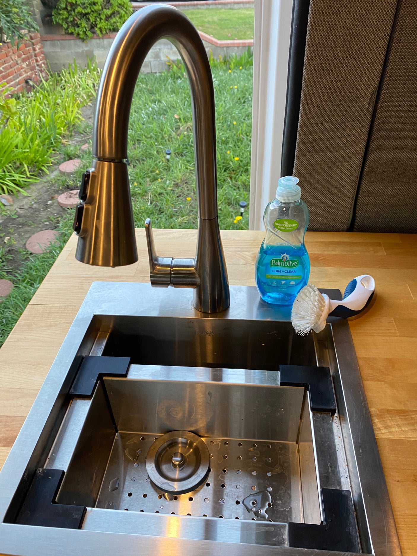 Compact RV rental kitchen sink with a modern faucet, Palmolive dish soap, and brush on a wood counter, overlooking greenery.