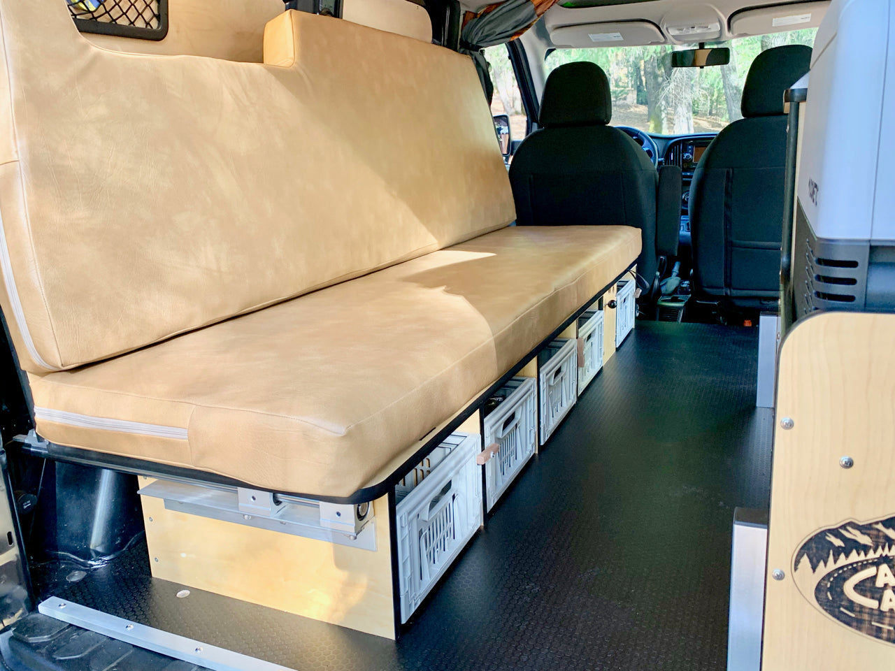RV rental: Camper van interior. Long beige bench seat, storage below, black floor, and front cab view visible.