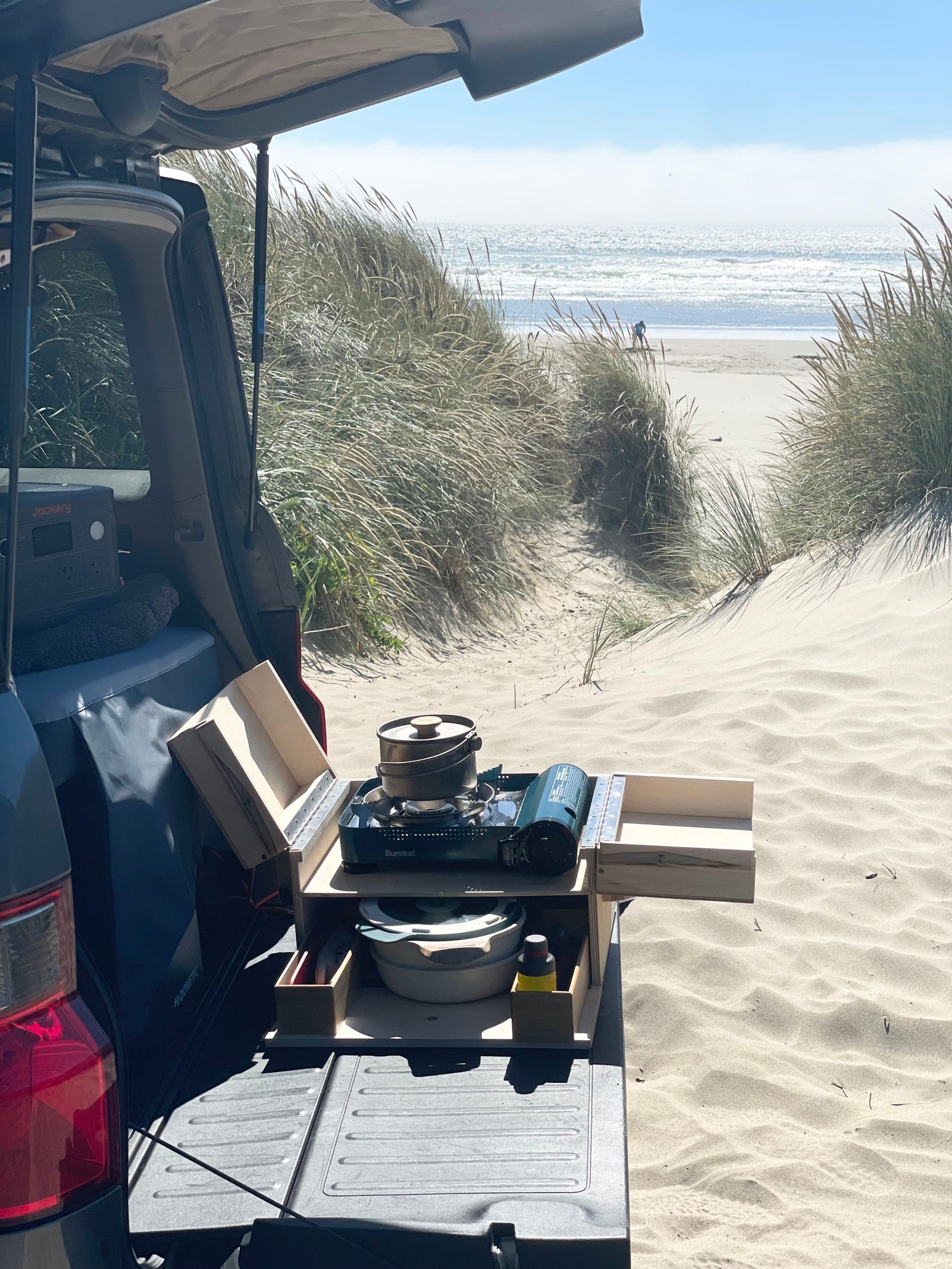 Cooking by the beach from a vehicle's tailgated setup. Features a portable stove, pots, and Jackery power, great for RV rental excursions.