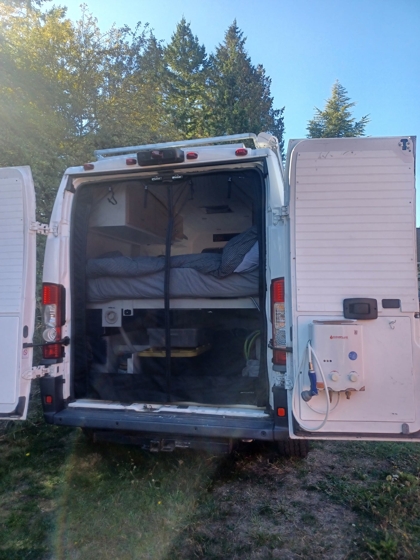 Open rear of an RV rental camper van revealing a cozy bed, storage, and an outdoor shower, set against trees & blue sky.