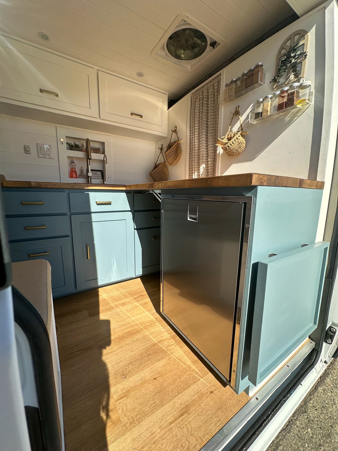 Bright RV rental kitchen with light blue cabinets, wood counters, fridge, and clever spice racks. Perfect for campervan adventures.