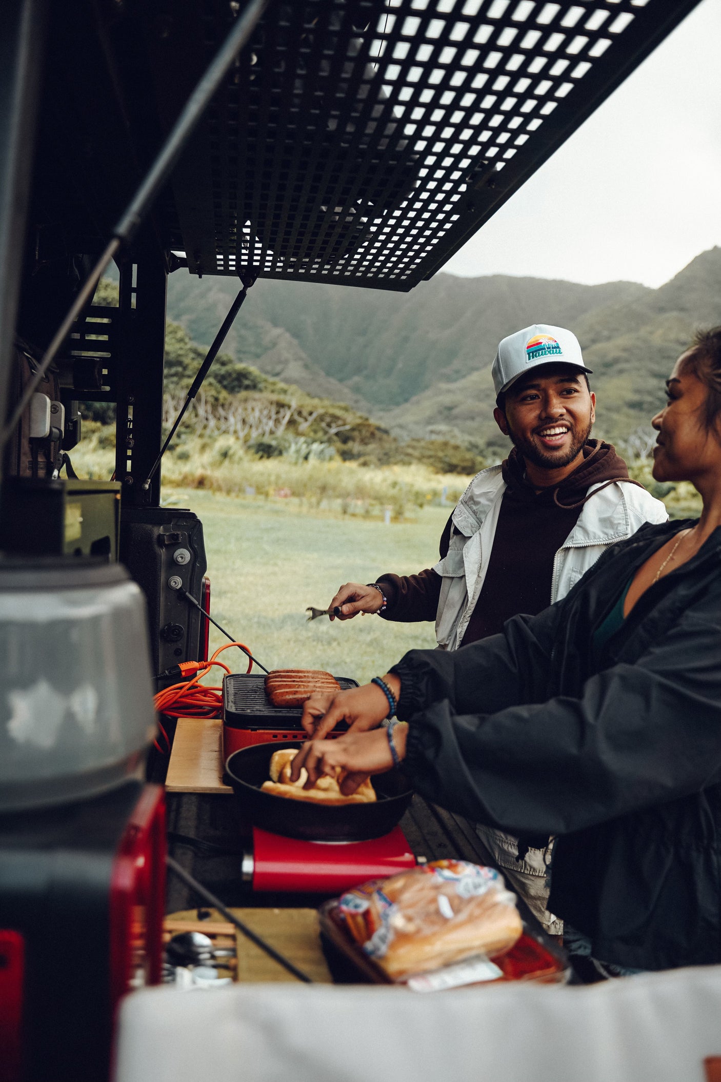 Enjoying an RV rental adventure! Two people cook sausages and bread from the vehicle's rear, with scenic mountains behind.