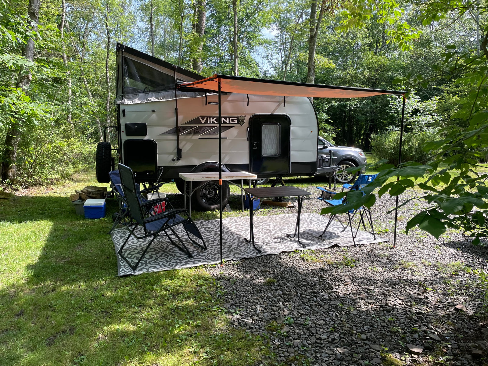 Viking pop-up RV rental comfortably set up at a wooded campsite, complete with awning, chairs, and tables for outdoor enjoyment.