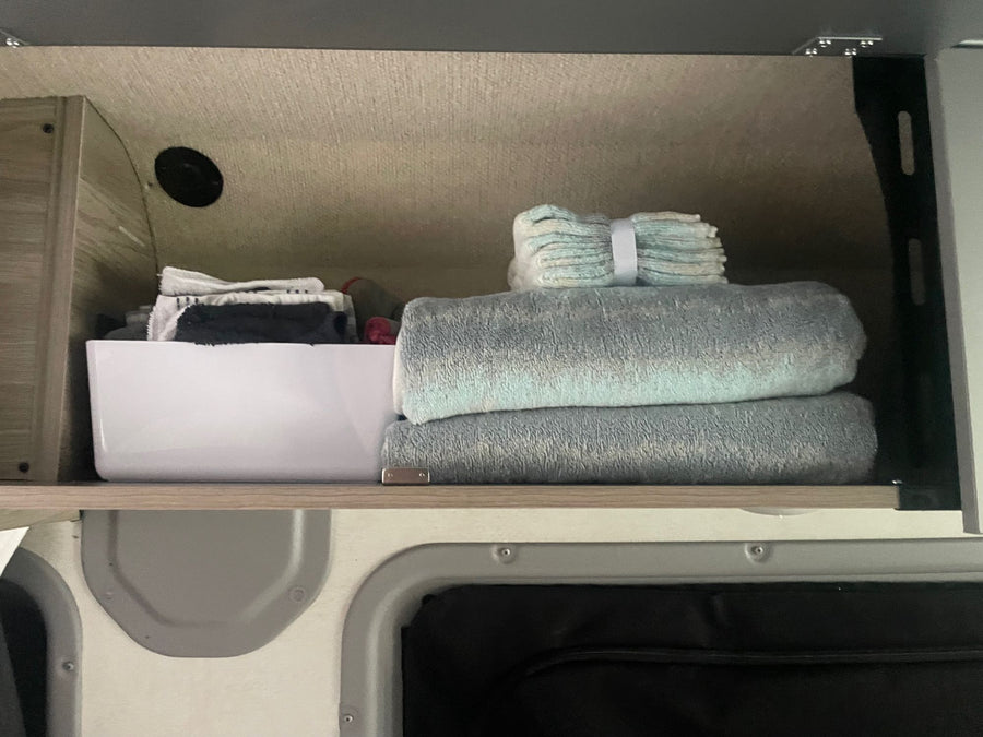 Inside an RV rental, an overhead cabinet reveals folded blue towels, smaller washcloths, and a white storage bin on a wooden shelf.